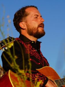 Dan Shukis, dressed as a cowboy, showcasing his musical talent by playing an acoustic guitar.