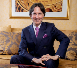 Dr. John Demartini, in a suit, gracefully seated on a couch, radiating sophistication and poise.