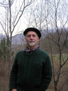 Dr. Brian Hayden, wearing a green sweater and hat, stands amidst the serene woods.