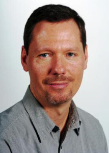 Dr. David Skrbina, in a grey shirt and tie, confidently gazes at the camera.