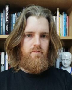 Dr. Jack Hunter, a bearded man with long hair, stands confidently before a bookcase.