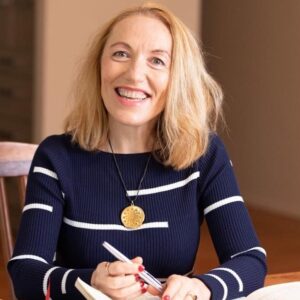 Dr. Joanna Kujawa, wearing a smile, sits at a table, engrossed in writing with a notebook and pen.