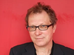 Portrait of Gary Lachman, wearing glasses and a black shirt, set against a striking red backdrop.