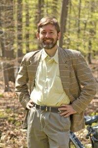 Andy Paquette in suit next to bike in woods.